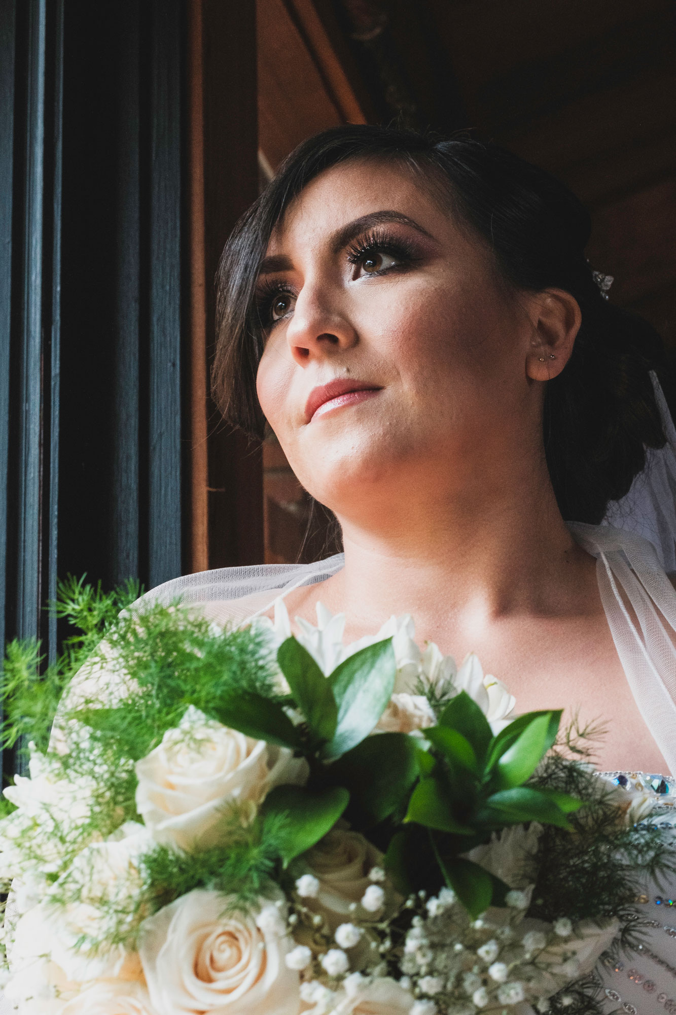 Karla beside a window in her wedding dress, holding a bouquet of white flowers.