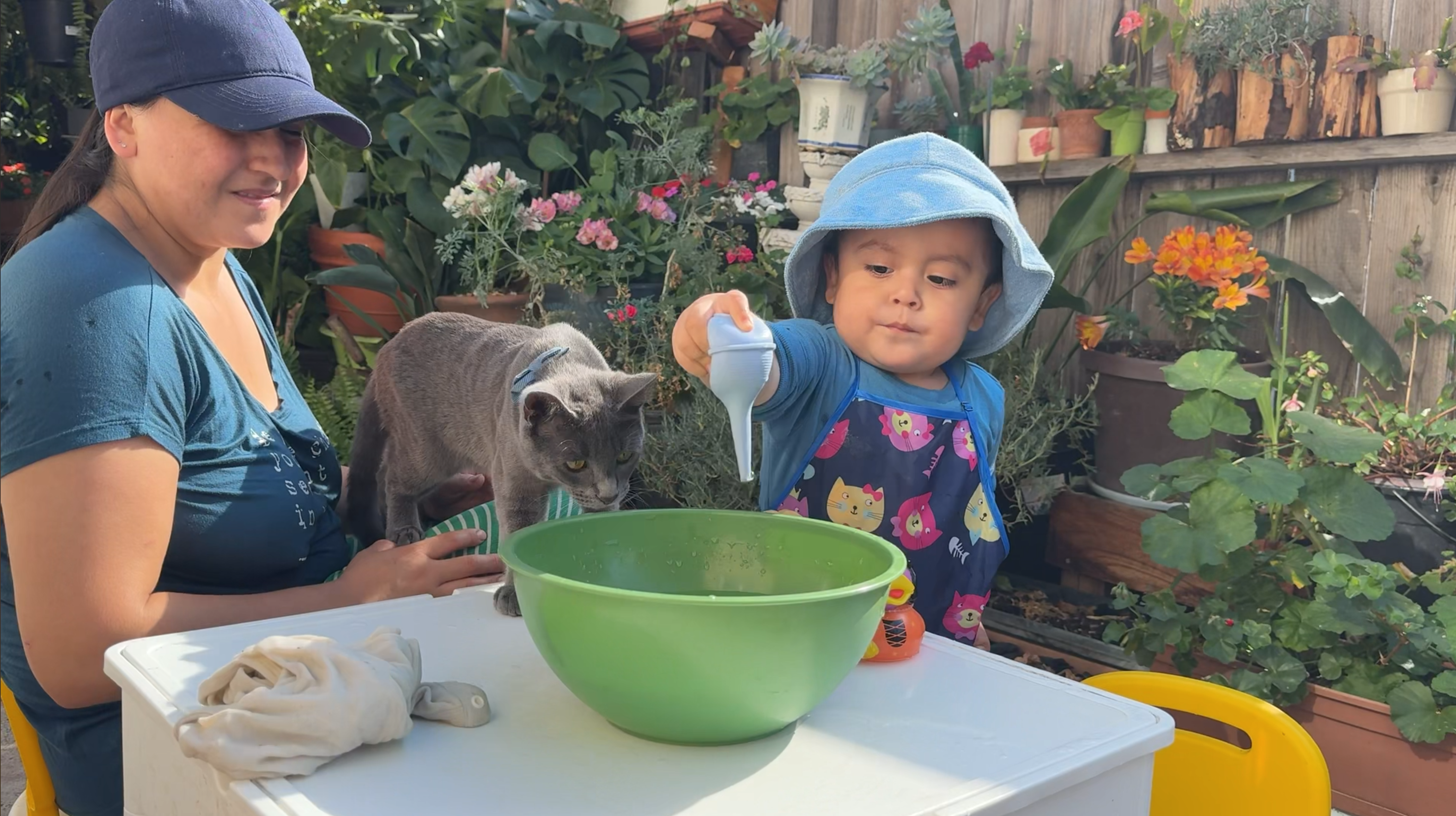 Karla and Gael playing with water in the garden with a gray cat beside them.